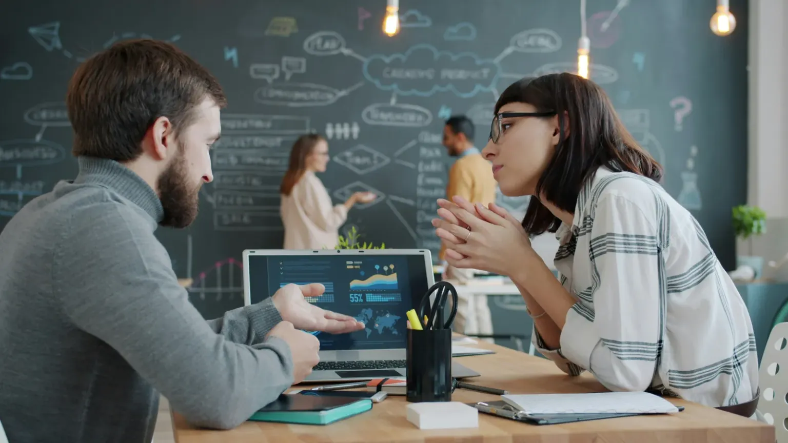 Two colleagues discussing charts on a laptop with a diagram-covered chalkboard behind them
