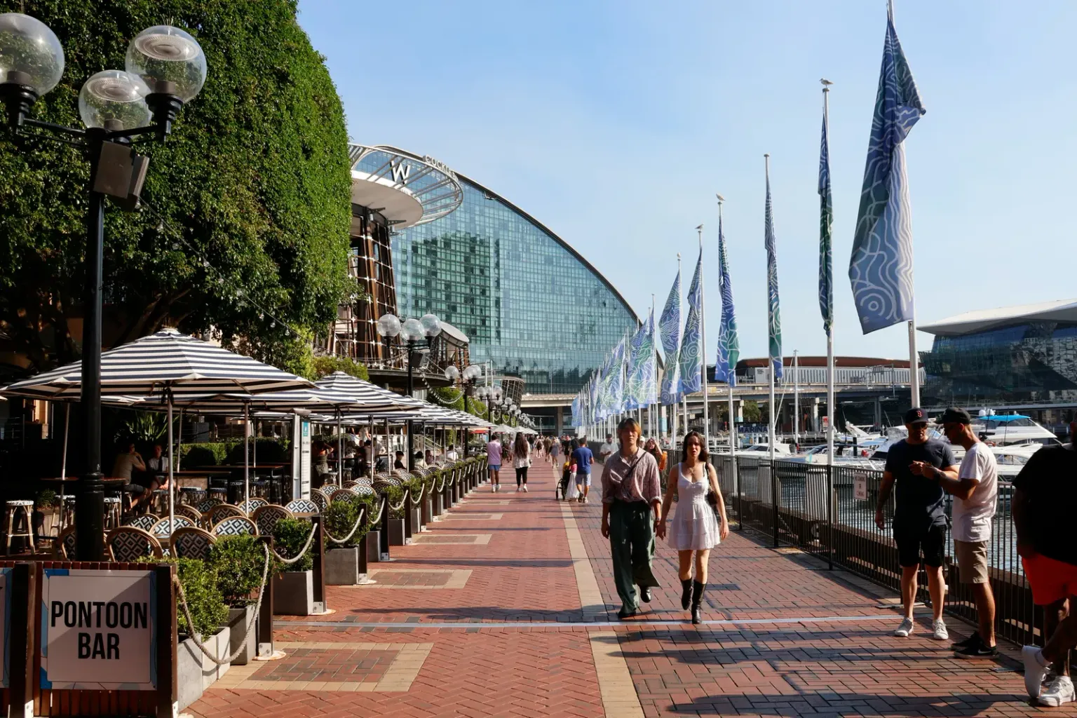 People walking along a waterfront promenade beside outdoor restaurants and a marina.